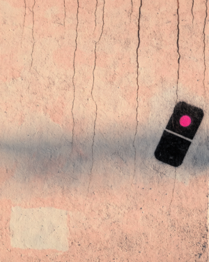 A small black domino stencilled near the edge of cracked, sun-bleached coral plaster, with one of its pips in hot pink.