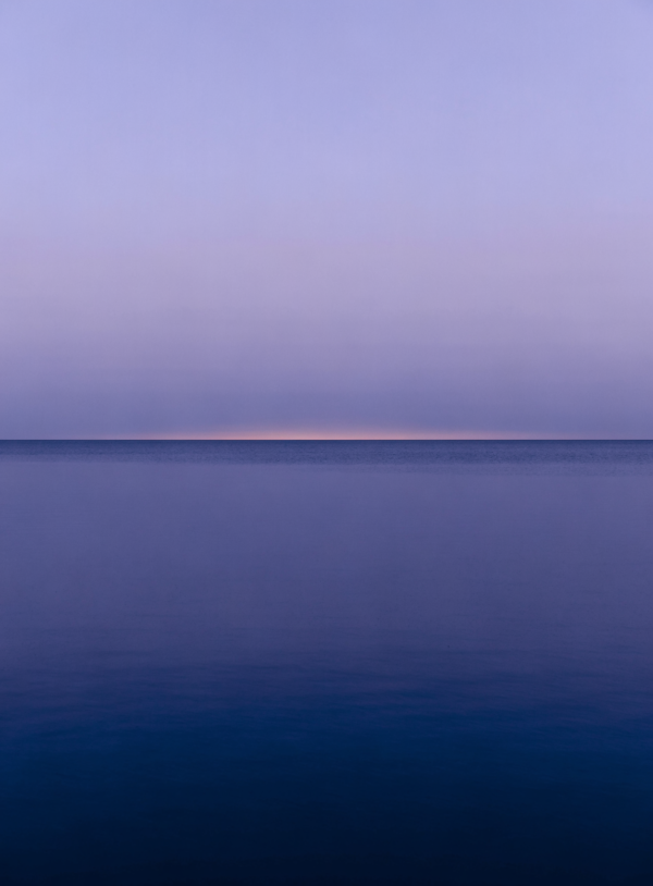A minimalist Nordic scene of a low, level horizon with pale gold light meeting cold blue above still, flat water and an empty sky.