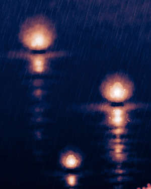 A tall indigo artwork of a rainy night with three soft warm lantern blooms, fine diagonal rain, a low drift of red maple at one edge and vertical light reflections on wet ground.