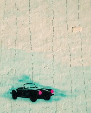 A stencilled convertible car with pink headlamps sprayed near the edge of a cracked mint, cream and coral plaster wall.