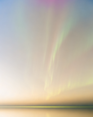 A flat low horizon of still water beneath a large sky of flowing green, magenta and cold blue aurora, with pale gold light along the waterline.